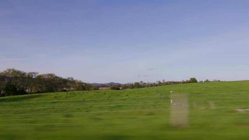 A side-view from a moving vehicle captures the rolling green hills and farms of the countryside on a sunny day