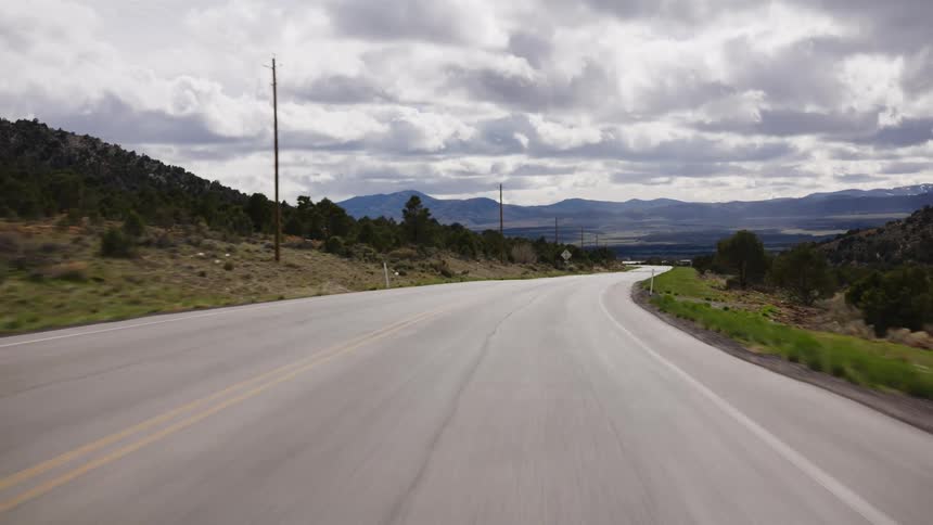 Point-of-view shot driving along a winding, rural two-lane highway through a mountain valley under a dramatic cloudy sky