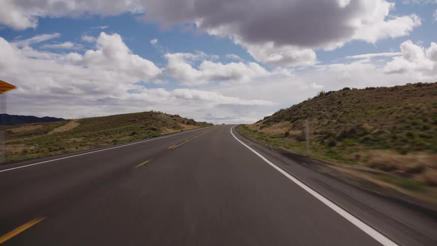 A first-person view of driving along a scenic two-lane highway through rolling hills under a cloudy blue sky