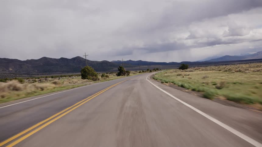 A point-of-view shot from a car driving down a rural highway in the American West with distant mountains under a cloudy sky
