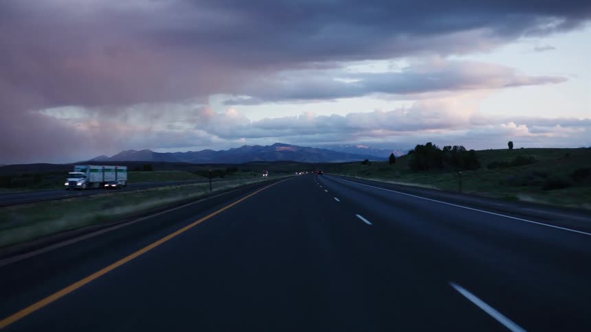 A point-of-view shot from a vehicle driving down a highway towards a mountain range under a dramatic, stormy sky at dusk
