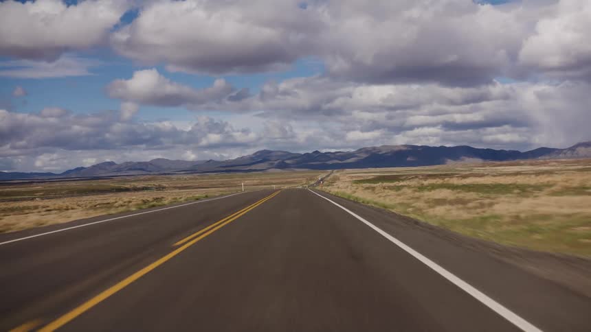 Traveling down an empty, straight highway through an arid landscape with mountains in the distance under a cloudy sky