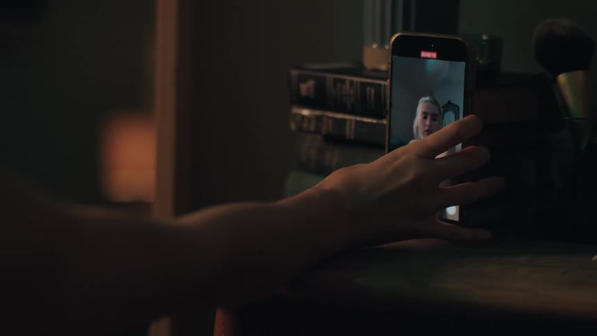 A woman's hand reaches out to touch her phone screen, which is recording a selfie video in a dimly lit room