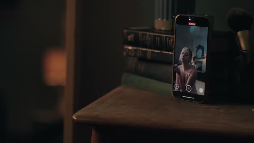 A smartphone on a wooden nightstand records a young woman applying makeup for a video tutorial