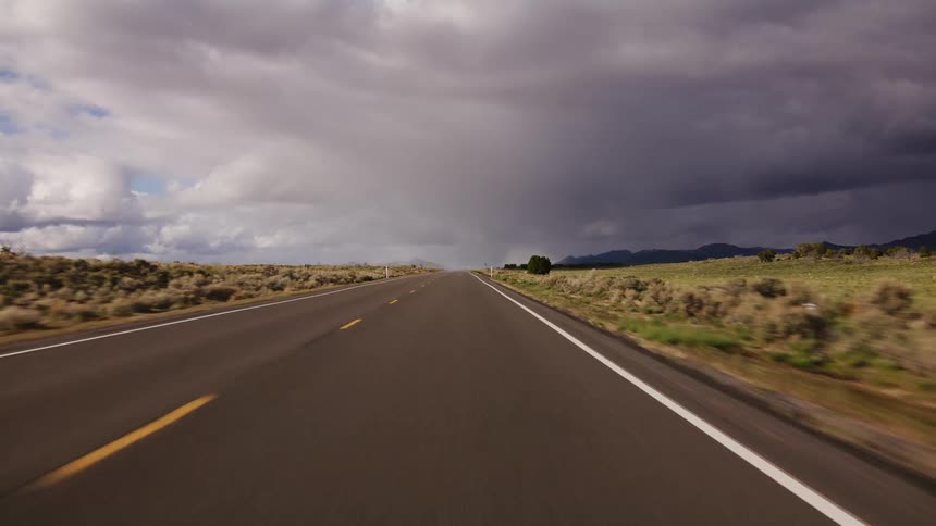 A point of view shot driving down a long, empty highway through a vast desert landscape towards a dramatic storm brewing in the distance