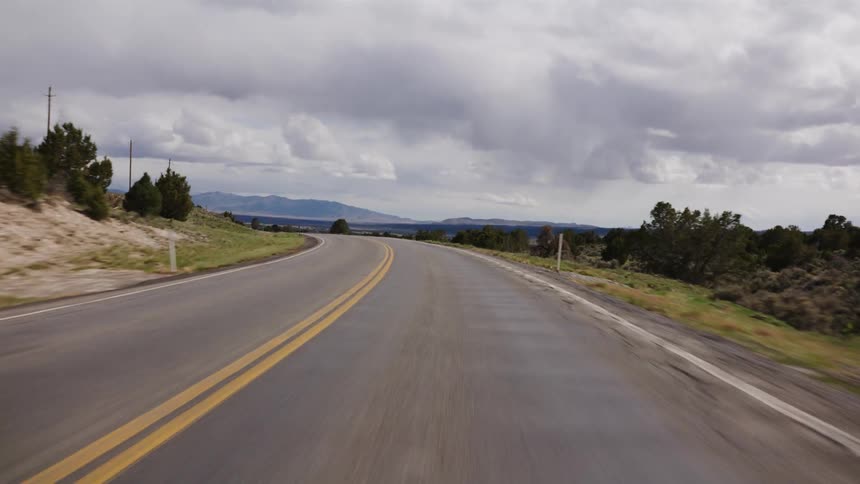 A point-of-view shot driving along a curving highway through a rural landscape with mountains in the distance under a cloudy sky