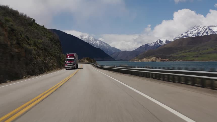 A scenic point-of-view drive along a winding mountain highway next to a beautiful reservoir with a majestic snow-capped mountain range in the background