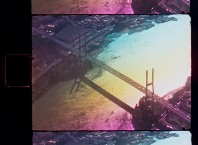 Inverted aerial view of traffic crossing a suspension bridge over a body of water captured on grainy, color-distorted vintage film
