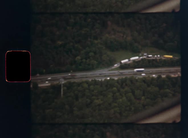 Super 8 film captures a distant, high-angle view of a major traffic backup on a highway interchange in a forested area