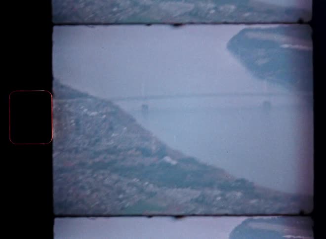 Vintage 8mm film captures a hazy, aerial view of a suspension bridge over a wide river