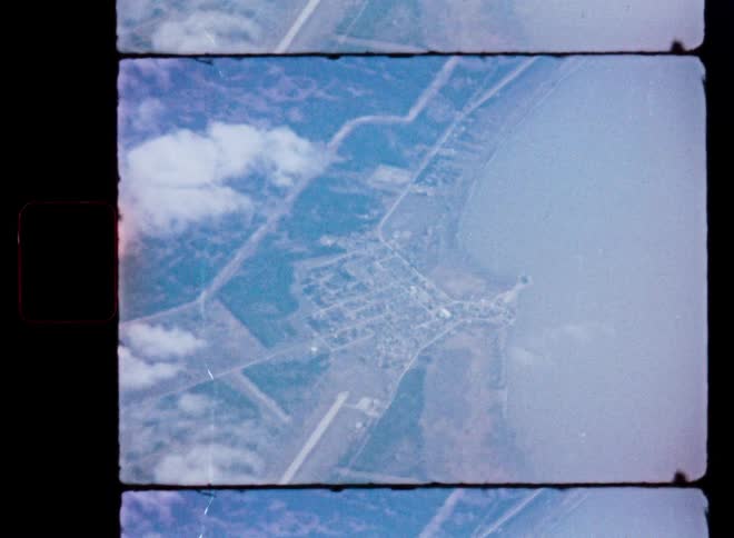 An archival high-altitude aerial view from 8mm film shows a small town and runway next to a large body of water