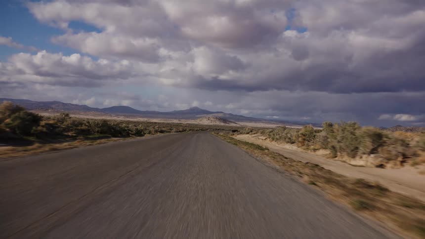 A driver's point-of-view traveling down an empty, winding desert road under a vast and dramatic cloudy sky