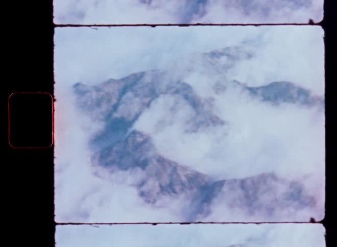 A vintage 8mm film scan shows a majestic mountain range peeking through a thick blanket of clouds from an aerial perspective