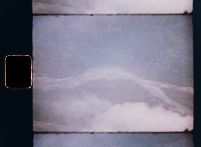 A vintage 8mm film captures an aerial view of a turquoise river winding through a mountainous forest landscape below the clouds