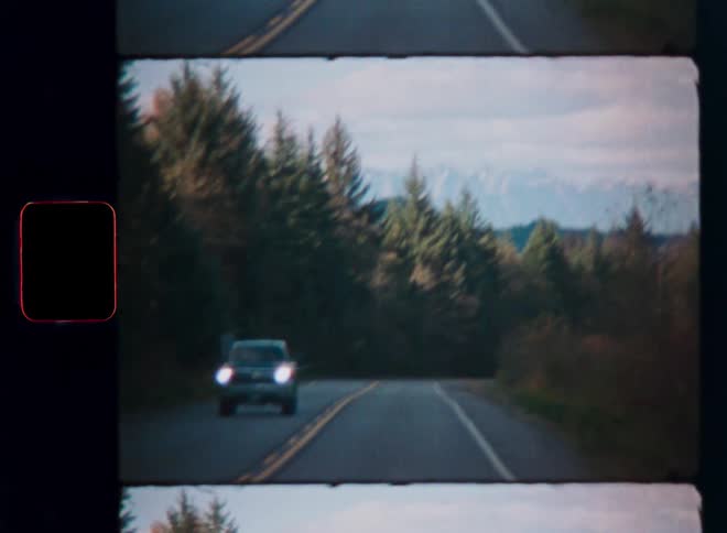Vintage 8mm film of a car driving on a rural road through a forest towards snow-capped mountains