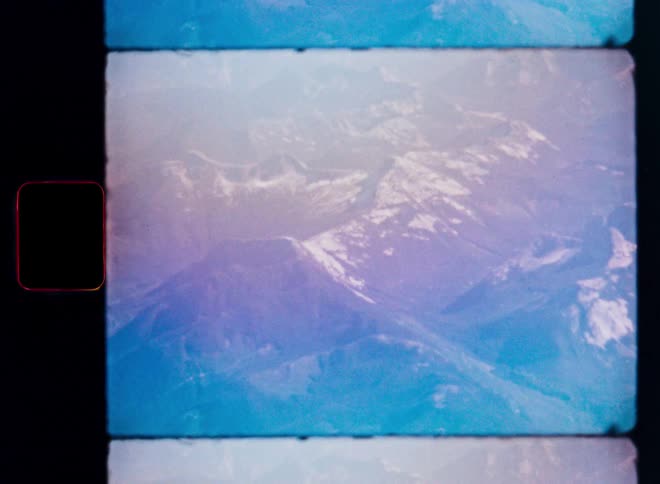 A dreamy, color-shifted vintage 8mm film captures a static aerial view of a snowy mountain range