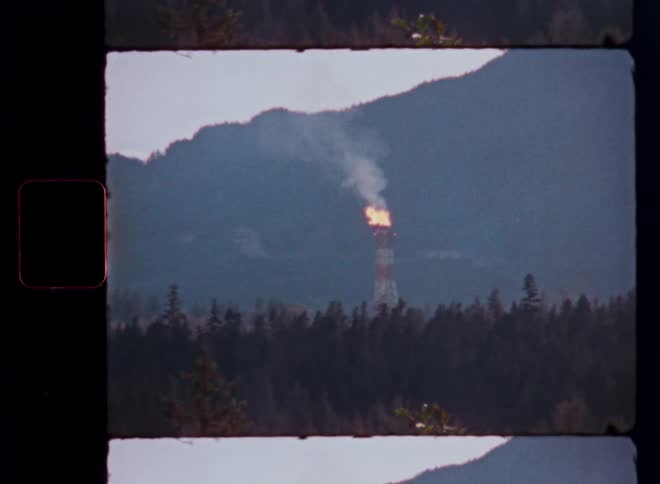 A distant industrial flare stack burns fire and smoke in a remote forested mountain landscape on vintage film