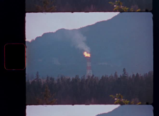 Vintage 16mm film footage of an industrial flare stack burning in a remote forest at the base of a mountain