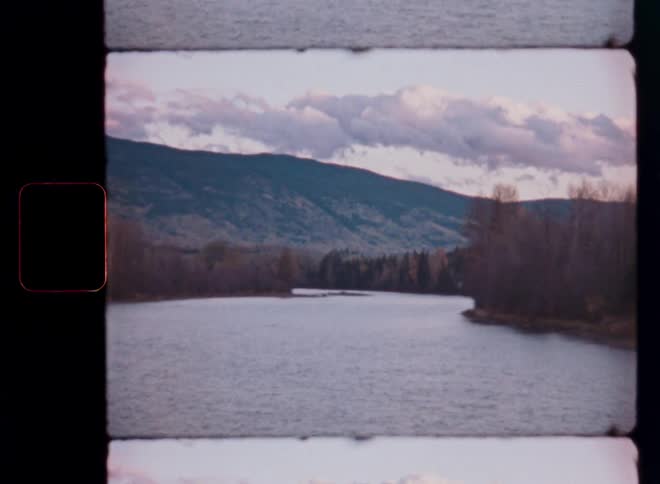 Vintage film captures a serene river landscape with forested mountains under a cloudy, atmospheric sky