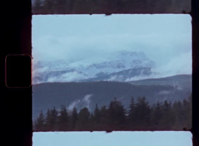 A scenic drive offers a view of snowy mountains and pine trees on a cloudy day, captured on vintage Super 8 film