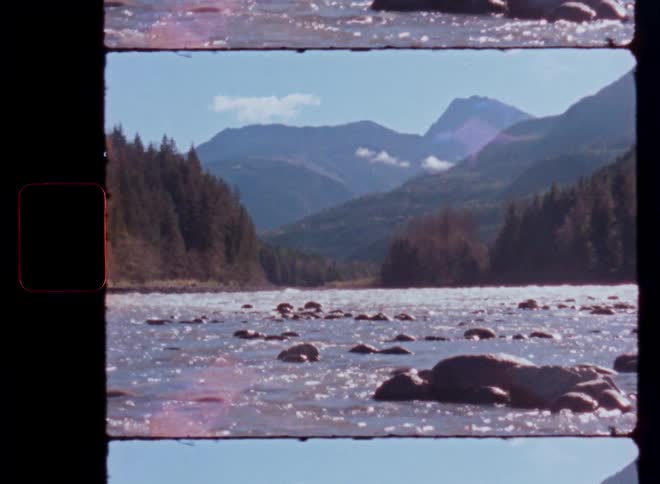 Vintage 16mm film captures a wide, sparkling river flowing through a forested mountain valley on a sunny day