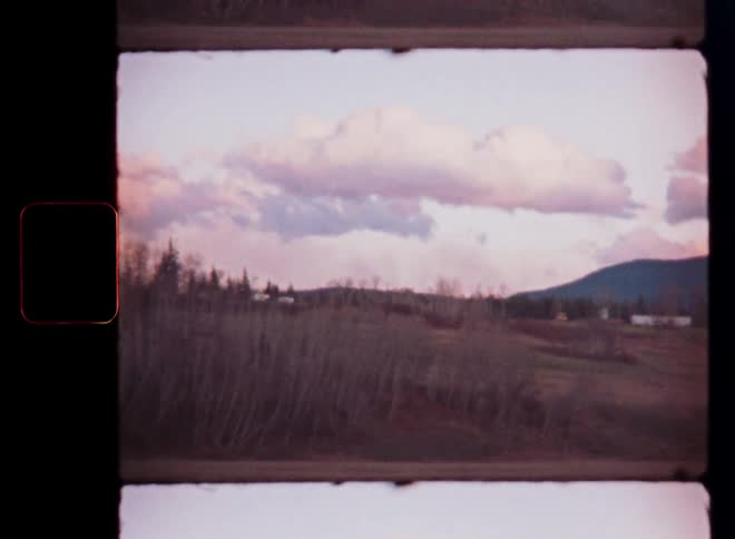 A nostalgic vintage film frame captures a tranquil countryside scene with rolling hills and bare trees under a pastel-colored cloudy sky