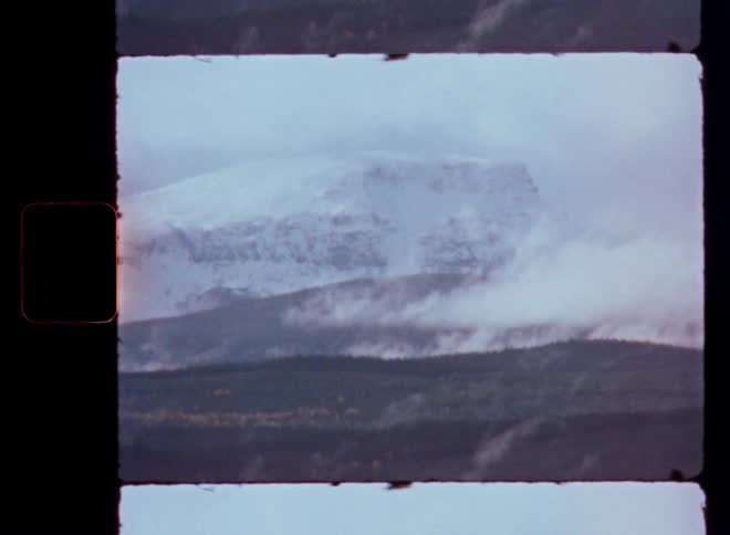 A vintage 16mm film scan captures a misty landscape of snow-capped mountains and rolling autumn hills