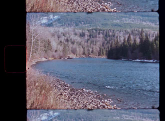 A vintage film scan captures a wide shot of a serene river flowing through a forested valley with snow-capped mountains in the distance