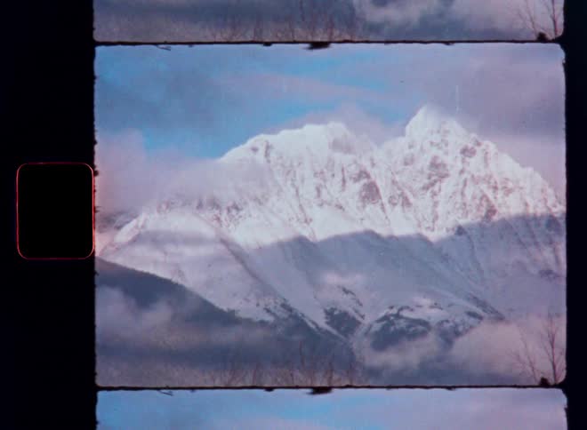A vintage film scan captures a static shot of a majestic, snow-covered mountain range with clouds in the sky
