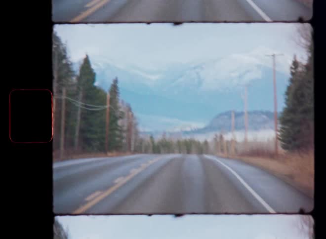 Handheld vintage film footage captures a view down a rural highway towards distant snowy mountains