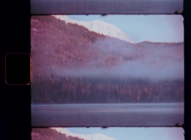 Vintage film footage captures a serene, misty lake reflecting a dense forest and distant snow-capped mountains