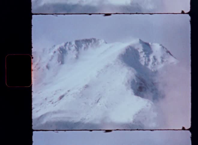 A vintage 8mm film reveals a majestic, snow-covered mountain peak emerging from the clouds