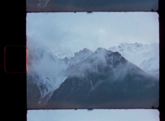 Point-of-view driving past majestic, snow-covered mountains on a cloudy day, captured on vintage film