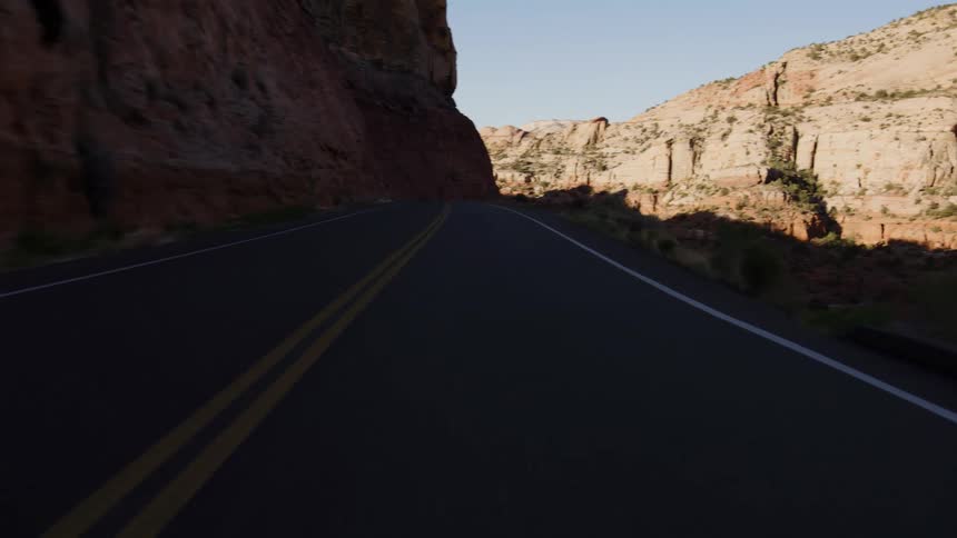 A point-of-view shot emerges from the darkness of a canyon shadow into the bright morning sun while driving along a winding desert highway