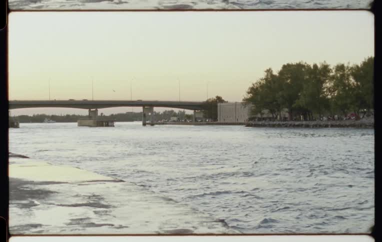 A serene, vintage film shot shows cars crossing a bridge over a calm waterway at dusk