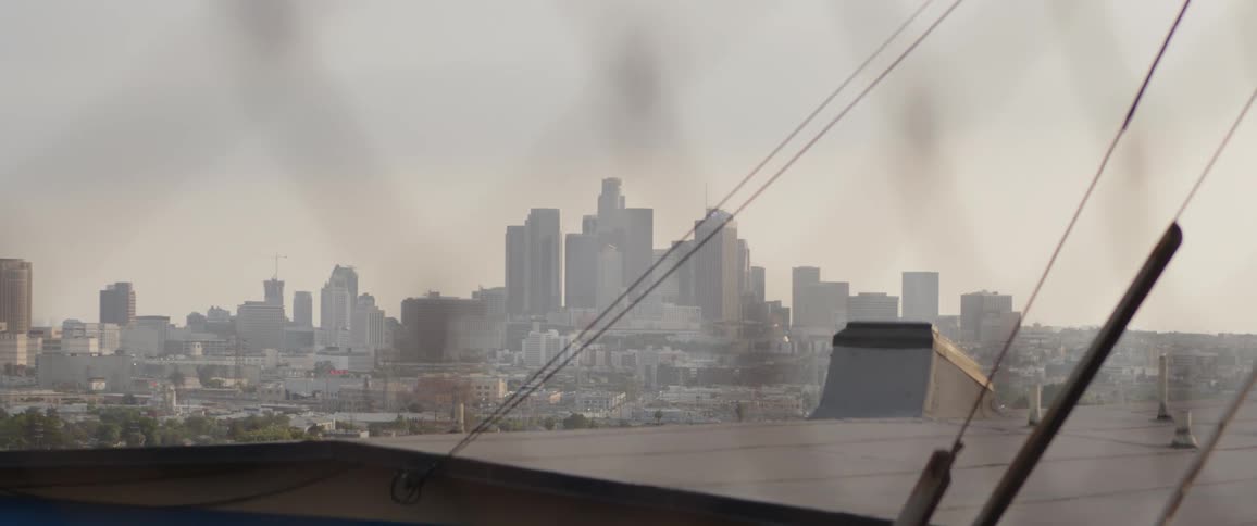 A rack focus shot slowly reveals the hazy downtown Los Angeles skyline behind a rusty chain-link fence