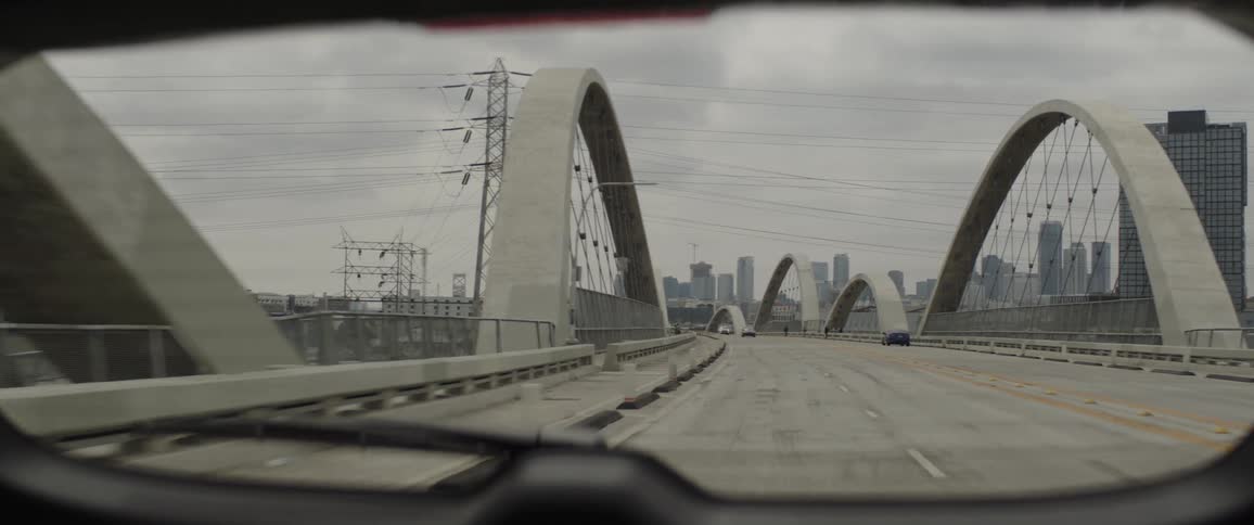 A point-of-view shot from a car driving across the 6th Street Viaduct towards the Los Angeles skyline on a cloudy day