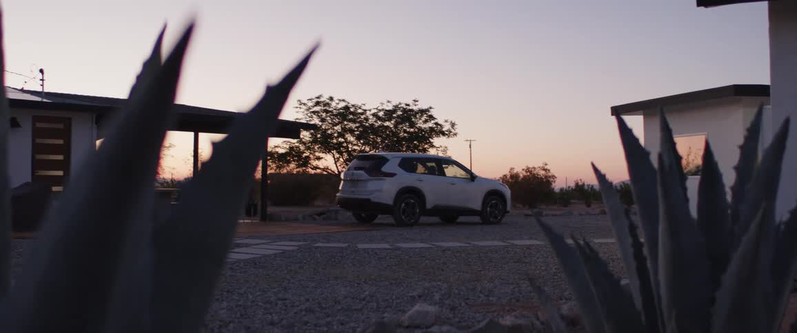 A white SUV is parked in the gravel driveway of a modern desert home at sunset, framed by agave plants