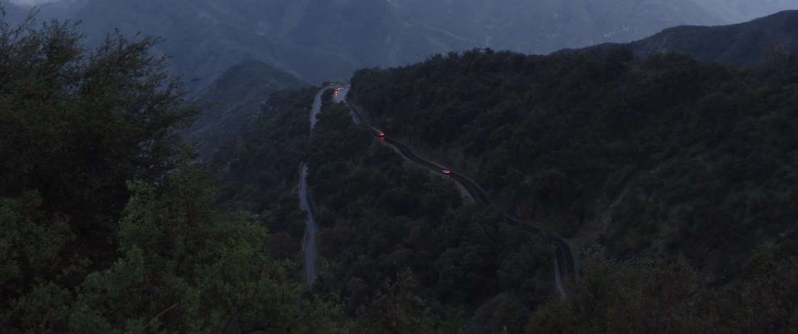 Traffic moves slowly down a winding mountain road through a dark, tree-covered landscape at dusk