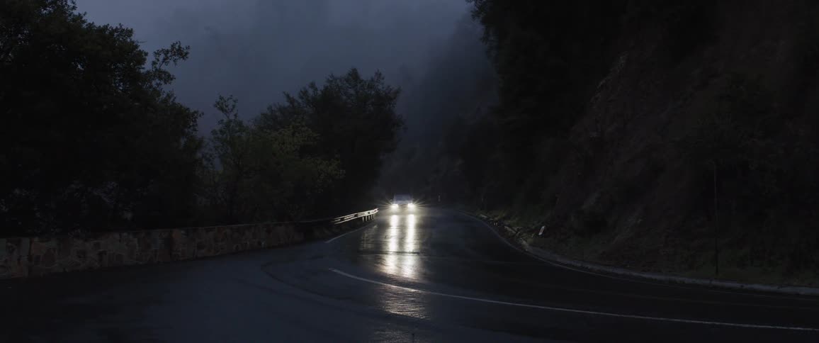 A white car drives around a bend on a dark, wet, and foggy mountain road