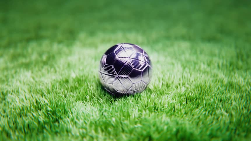 A surreal, spinning dolly-in shot focuses on a silver metallic soccer ball resting on vibrant green grass