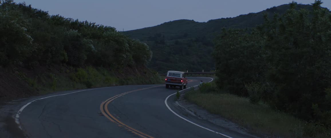 A vintage red SUV drives away down a winding, remote mountain road at dusk