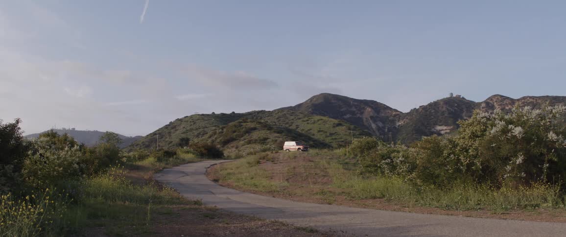 A wide shot shows a white van parked on a grassy hill in a lush, mountainous landscape with a winding road in the foreground