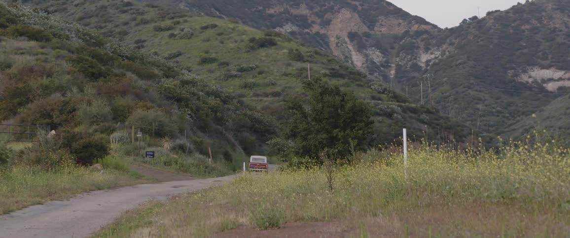 A vintage orange SUV drives away down a narrow country road that winds through lush green mountains