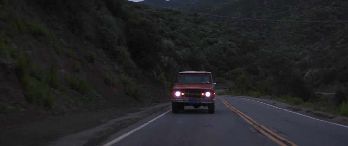 A vintage red SUV with headlights on drives along a winding mountain road at dusk