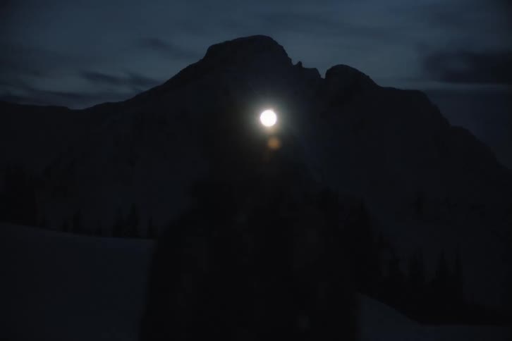 A silhouetted person wearing a headlamp in a dark, snowy mountain landscape brightens their light, creating a large lens flare
