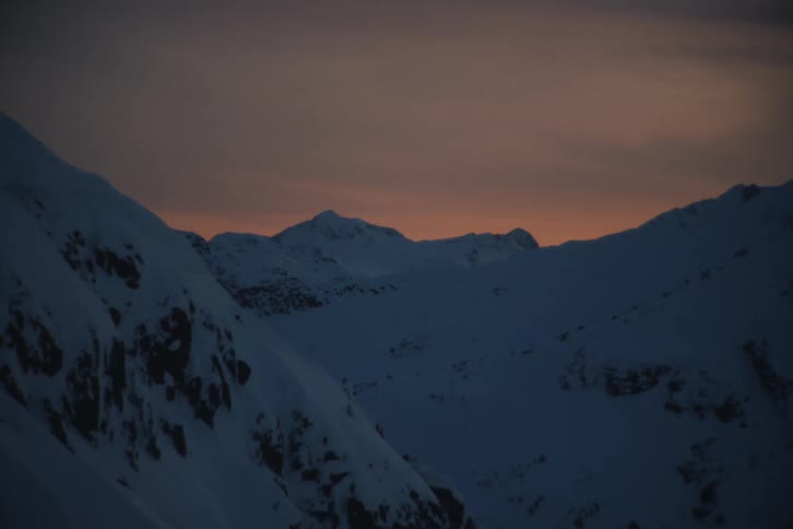 The last light of sunset casts a warm orange glow over a vast, snow-covered mountain range at dusk