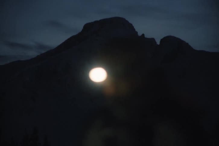 A bright light from a headlamp creates a lens flare as it moves across a silhouetted mountain at night