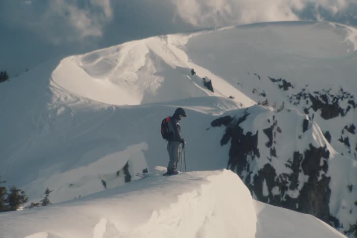 A fast zoom shot reveals a lone skier standing on the precipice of a vast, snowy mountain peak under a dramatic cloudy sky