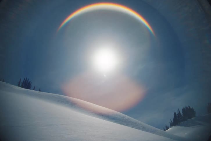 An atmospheric optical phenomenon creates a vibrant rainbow halo around the sun above a serene, snowy winter landscape